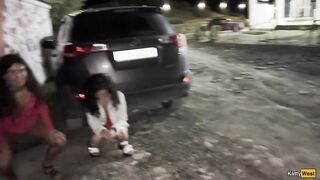 Two girlfriends pee together near a car in a public parking lot